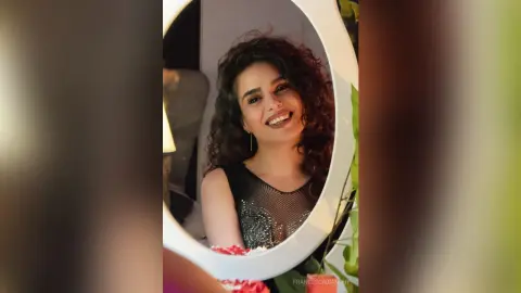 Smiling woman with curly hair in a black mesh top posing for a mirror selfie in warm indoor lighting.