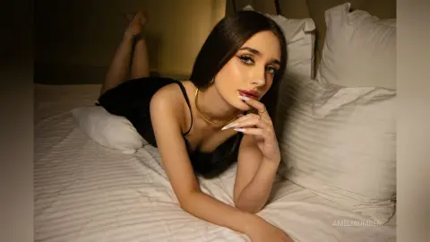 Young woman in a black tank top relaxing on a white bed in a moody, dimly lit bedroom setting.