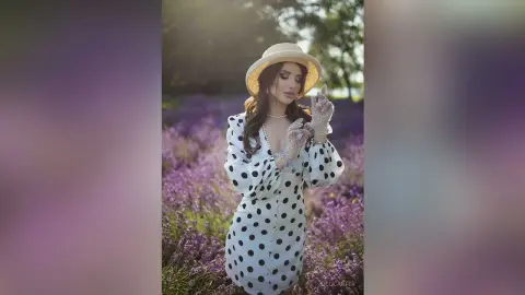 Elegant woman in a white polka dot dress and straw hat posing in a dreamy lavender field at sunset.