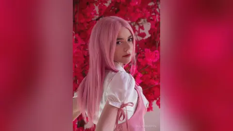 Young woman with long pink hair in a white blouse posing amidst vibrant pink flowers for a dreamy aesthetic.