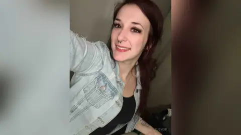 Smiling woman with red hair wearing a light denim jacket in a casual indoor setting.