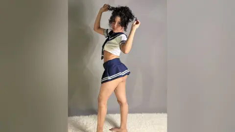 AkiraHayami poses in a white and navy sailor-style school uniform, showing her midriff and legs against a plain grey wall.