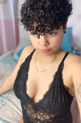 Young woman with curly dark hair wearing a black lace camisole and silver necklace in a bright bedroom.