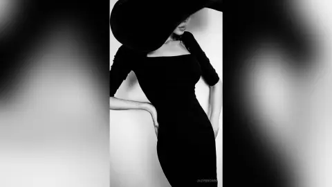 Elegant woman in a black cocktail dress and wide-brimmed hat posing in a minimalist studio setting.