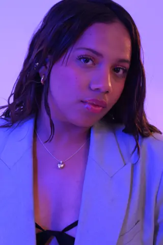 Young woman in a light blue blazer and heart necklace posing under moody purple studio lighting.
