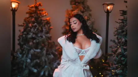 Elegant woman in a white faux fur robe posing amidst festive Christmas trees and warm glowing street lamps.