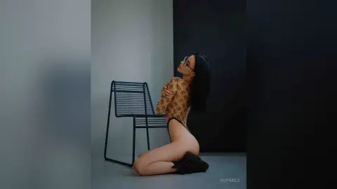 SofiMils poses kneeling on the floor in a patterned brown bodysuit, arching her back and looking up while wearing glasses.