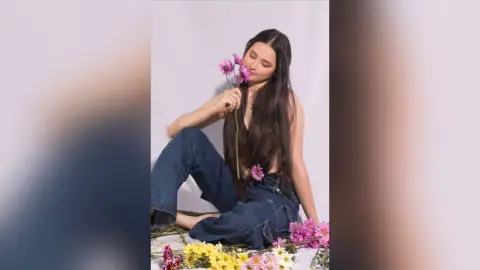 Young woman in denim jeans sitting against a white background holding pink flowers with a serene mood.