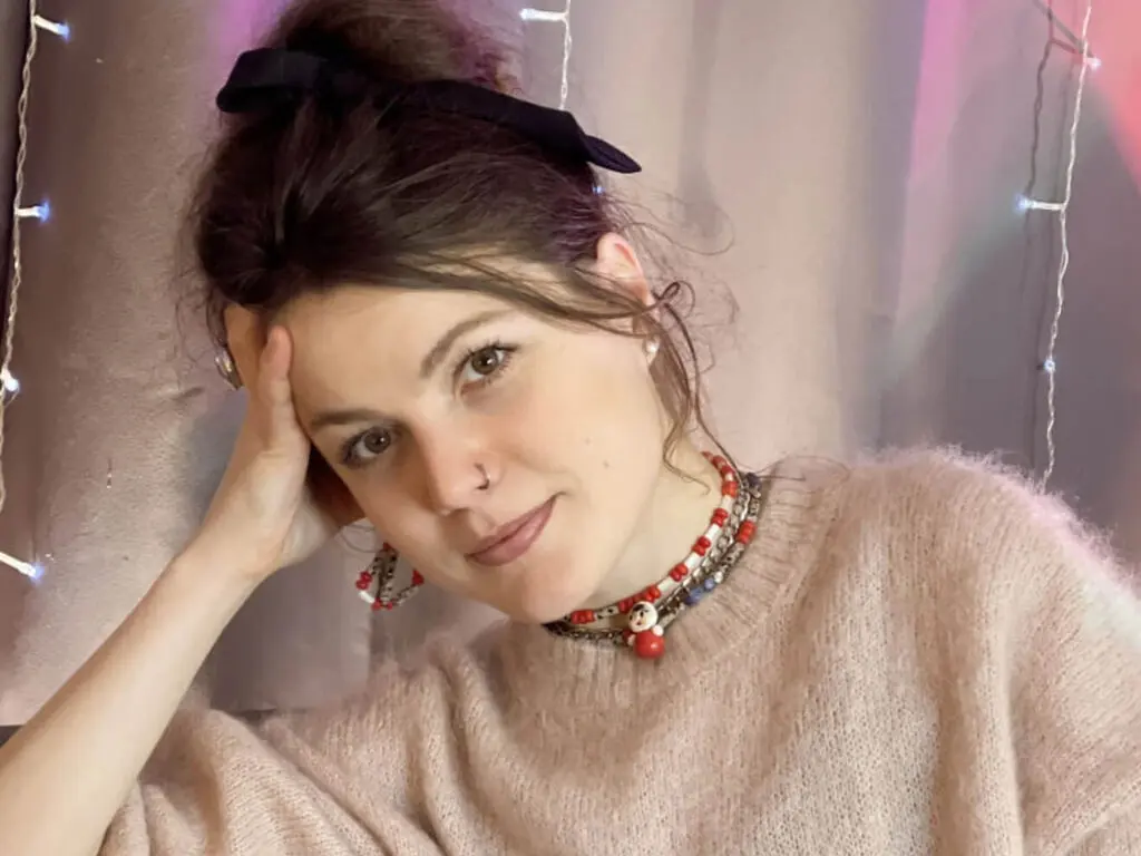 MarlaBlum poses with her hand against her cheek, wearing a light pink fuzzy sweater, a black hair bow, and a red and white beaded necklace.