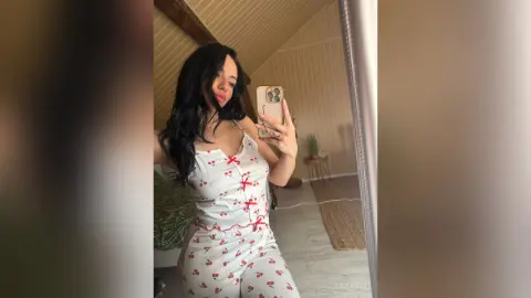 Young woman with long dark hair wearing a white floral print camisole in a bright, cozy bedroom setting.