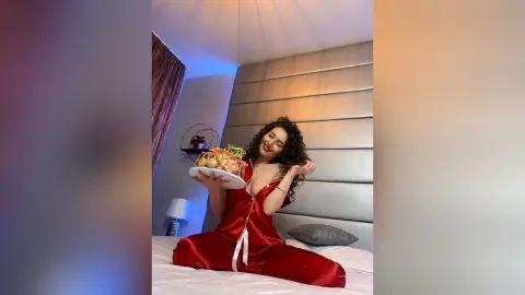 Smiling woman in red silk pajamas holding a celebratory dessert platter in a cozy bedroom setting.