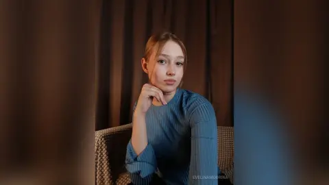 Young woman in a blue ribbed long-sleeve top sitting thoughtfully in a dimly lit room.