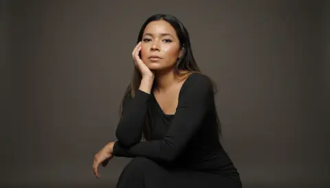 XinemaBonet poses with her hand against her face, wearing a long-sleeved black dress against a plain dark background.