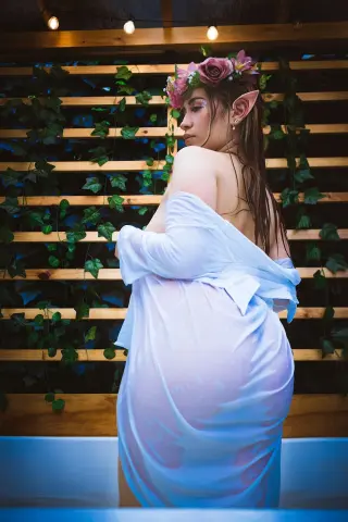 Ethereal elf woman in a white sheer dress wearing a flower crown in a garden setting.