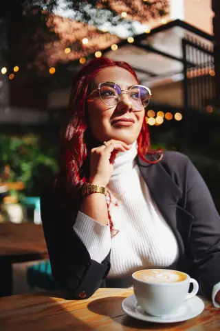 Thoughtful woman in a white turtleneck and black blazer enjoying coffee at an outdoor cafe at sunset.