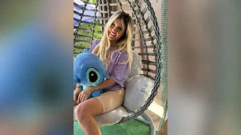 Anita smiles while sitting in a hanging egg chair, holding a blue Stitch plush toy and wearing a light purple t-shirt and tan shorts.