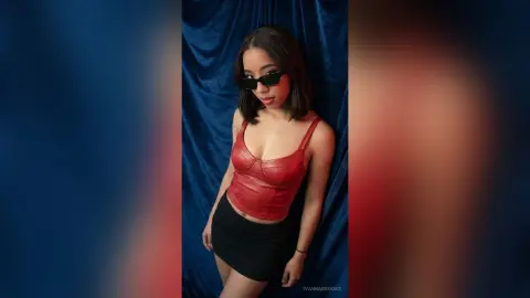 Young woman in a red corset top and black skirt posing against a blue velvet curtain backdrop.