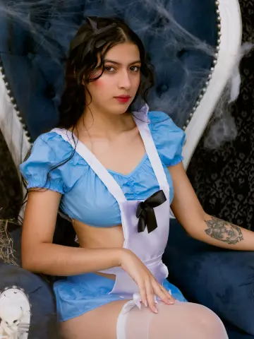 SusanaSanchez20 wears a light blue maid outfit with a white apron and black bow, sitting in a dark blue velvet chair surrounded by spiderwebs.