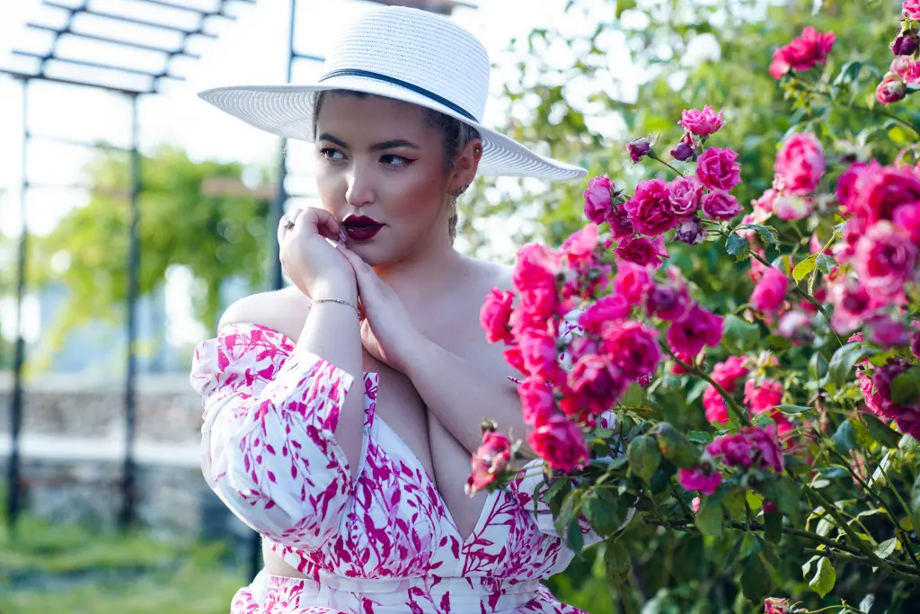 CleoLangley — Elegant woman in a white sun hat and pink floral dress posing amidst blooming pink roses in a sunny garden.