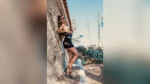 EvelynMills poses against a stone wall wearing a black bodysuit and sunglasses.