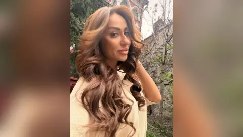 Smiling woman with long wavy brown hair wearing a beige knit sweater in a lush outdoor garden setting.
