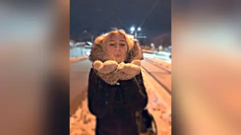 Blonde woman in a cozy fur-lined winter coat blowing a kiss on a snowy night street.