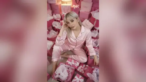 Blonde woman in pink silk pajamas sitting amidst luxury gift boxes in a dreamy pink aesthetic setting.