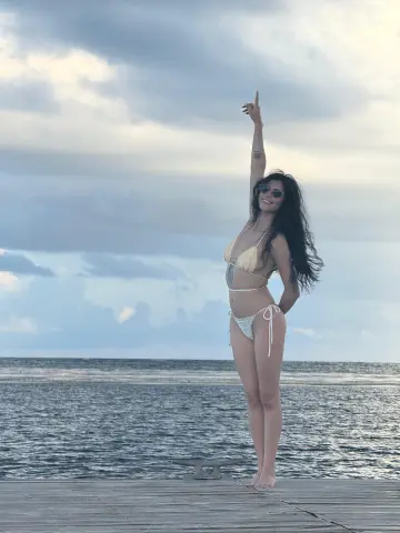Smiling woman in a white bikini posing on a wooden pier against a cloudy ocean backdrop.