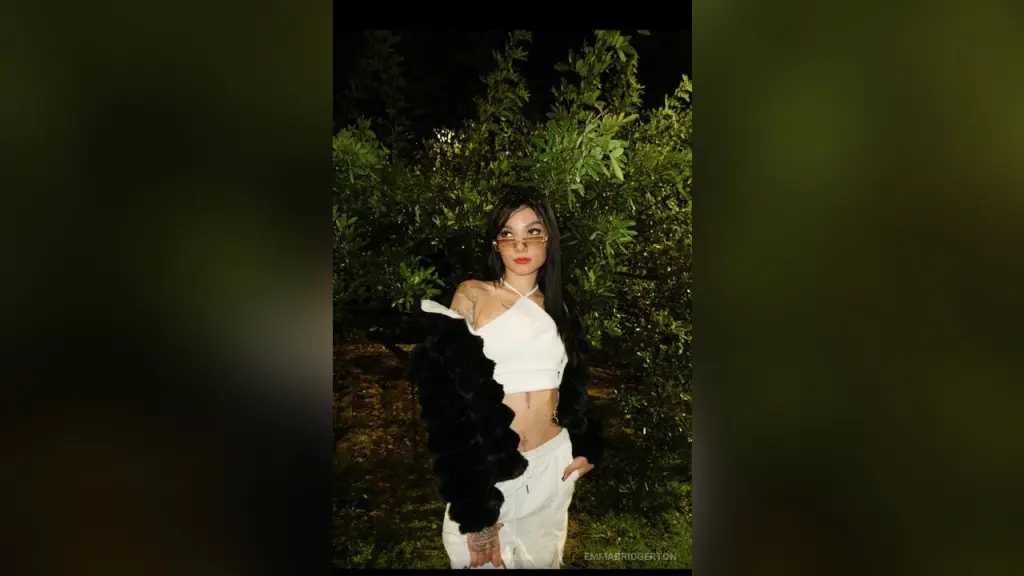 Young woman in a white halter top and black faux fur coat posing against lush green foliage at night.