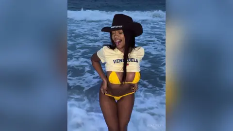 Smiling woman in a yellow Venezuela graphic tee and bikini posing on a sunny beach by the ocean waves.
