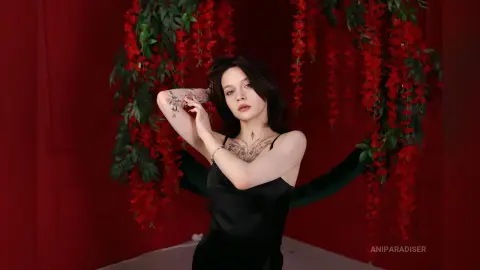 Tattooed woman in a black satin dress posing against a romantic red backdrop with hanging floral vines.