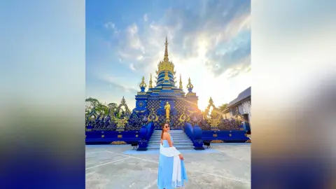 Woman in a light blue dress and white shawl standing before a majestic blue Thai temple under a sunset sky.