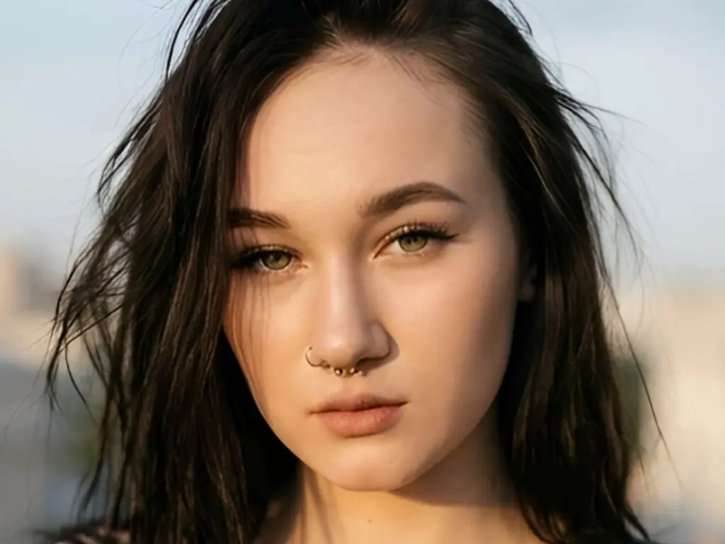 EllaVietor looks directly at the camera with long, dark hair and a septum piercing, set against a soft, outdoor background.