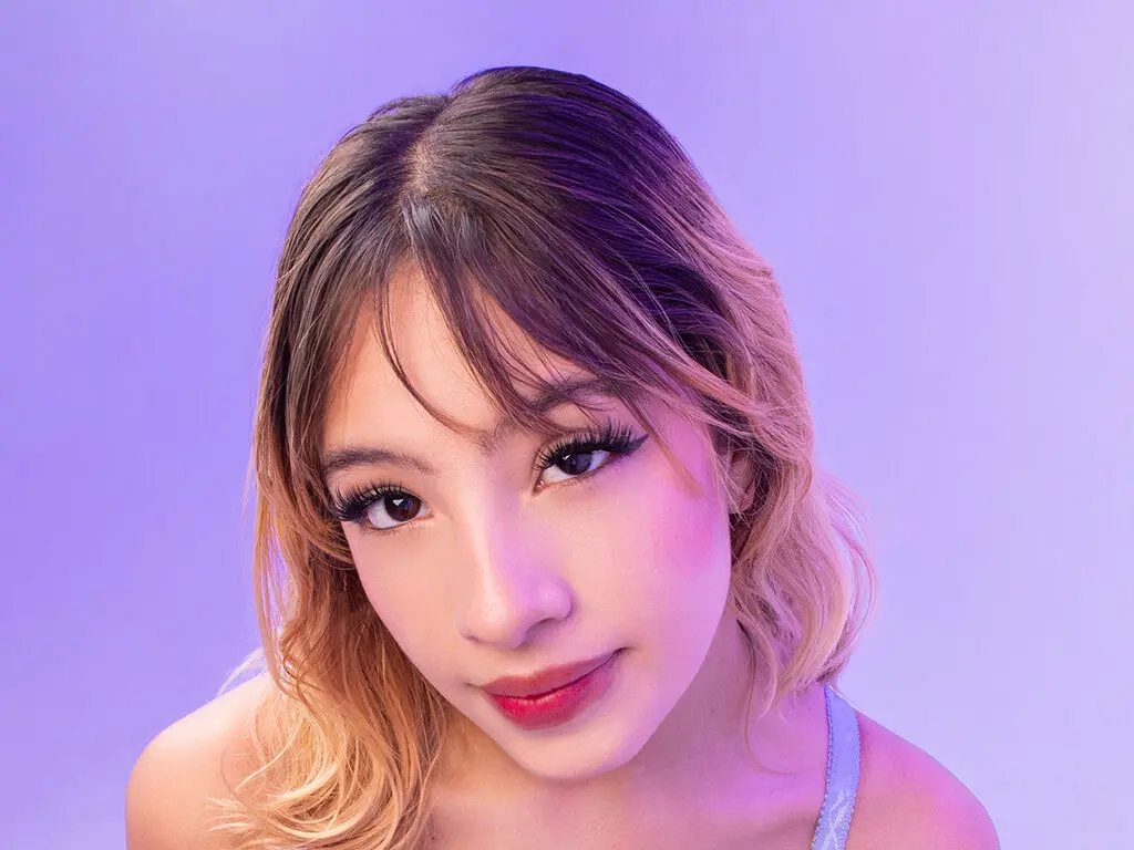 Young woman with blonde hair and dramatic eyelashes posing against a soft purple background.