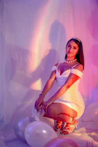Young woman in a white cocktail dress and pearl necklace posing against a dreamy pink and purple backlit background.