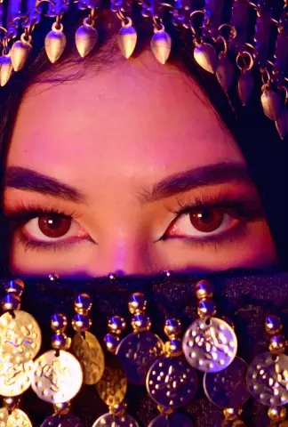 Close-up of a woman with dramatic eye makeup wearing a traditional gold coin headpiece in warm, moody lighting.