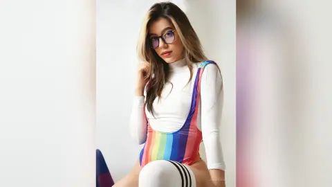 JessieLiu wears a rainbow-striped bodysuit and glasses while posing against a white background.