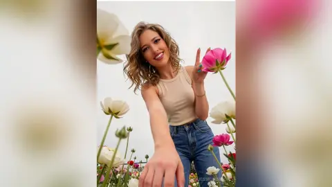 Smiling young woman in a beige tank top and blue jeans posing amidst white and pink flowers in bright daylight.