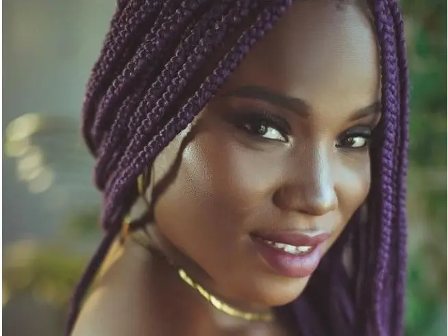 Smiling Black woman with long purple micro braids and gold jewelry in a soft outdoor setting.