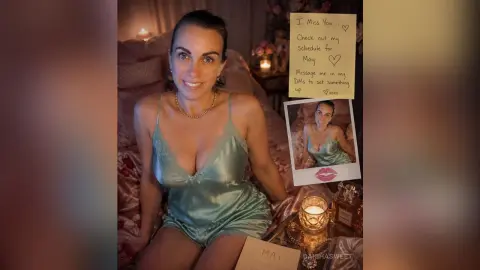 Smiling woman in a light blue satin camisole sitting in a cozy, candlelit bedroom setting.