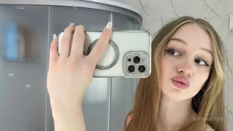 Young woman with long blonde hair taking a selfie in a mirror with a white phone case in a bright room.