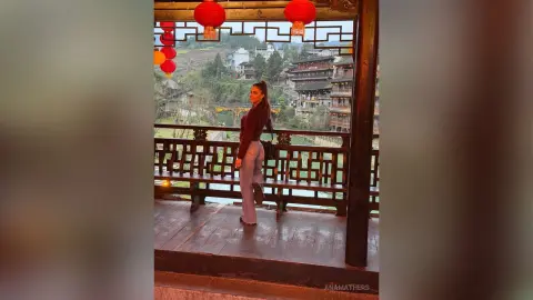 Woman in casual outfit standing on a traditional wooden balcony with red lanterns overlooking a scenic village.