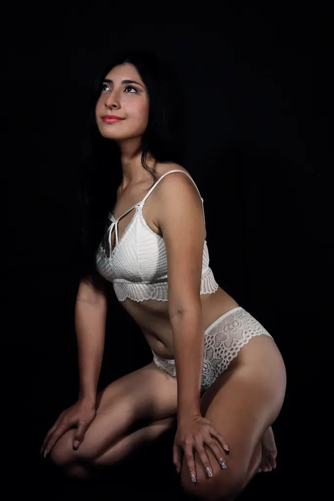 EmmaWilliaamss poses in a black lace outfit with sheer mesh sleeves and a dark skirt, looking over her shoulder against a plain light background.