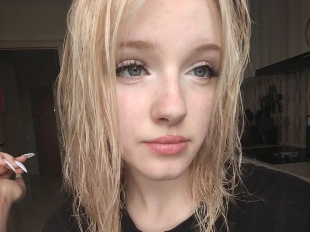 Close-up portrait of a blonde woman with wet hair looking thoughtfully at the camera in a dimly lit room.