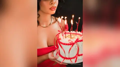 Glamorous woman in a red dress holding a white birthday cake with lit candles in a moody, celebratory setting.