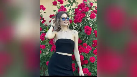 ElinaCarter poses in a black strapless top and skirt, wearing sunglasses and a necklace, standing in front of a bush of bright red roses.