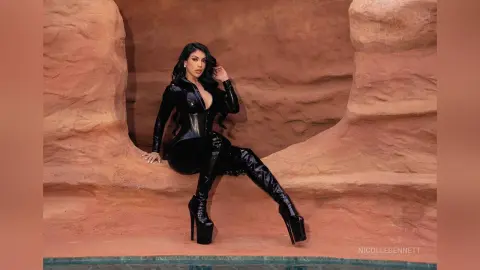 Glamorous woman in a black latex bodysuit and thigh-high platform boots posing in a desert canyon setting.