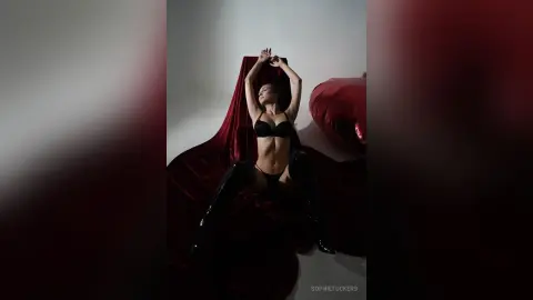 Slim woman in black lingerie and thigh-high boots posing against red velvet fabric in a moody studio setting.