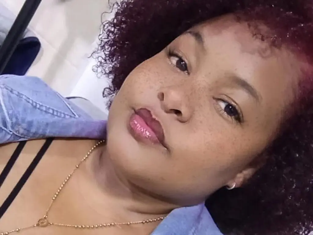 Close-up portrait of a woman with curly reddish hair wearing a denim shirt and gold necklace in soft lighting.