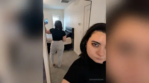 Young woman in a black t-shirt and grey trousers taking a mirror selfie in a bright, modern bedroom.